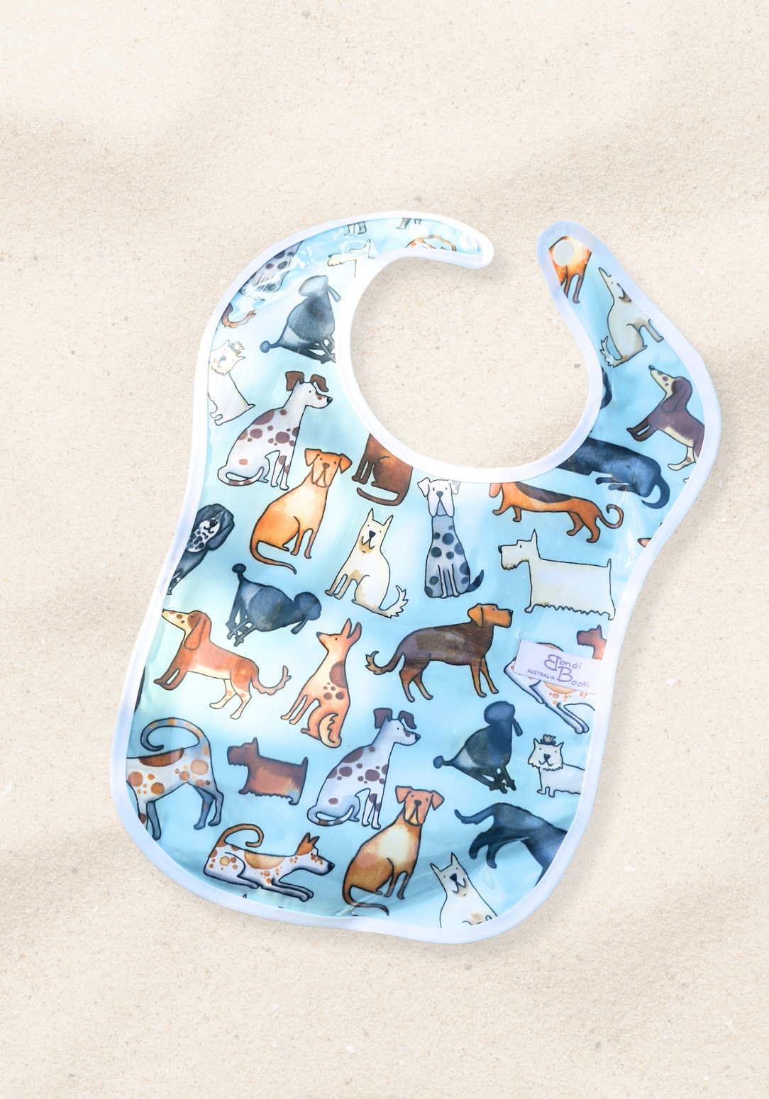 Laminated baby feeding bib featuring a playful pattern of illustrated dogs in various breeds and colors on a light blue background, trimmed in white with a snap closure, laid flat on a sand background.