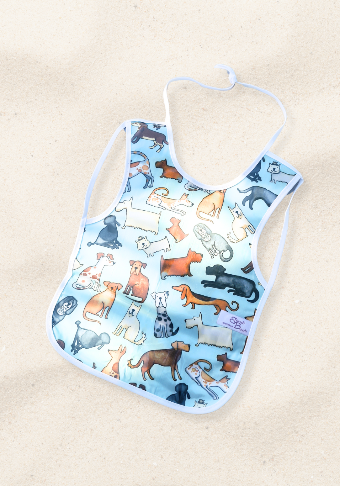 Laminated baby feeding bib featuring a playful pattern of illustrated dogs in various breeds and colors on a light blue background, trimmed in white with a snap closure, laid flat on a sand background.
