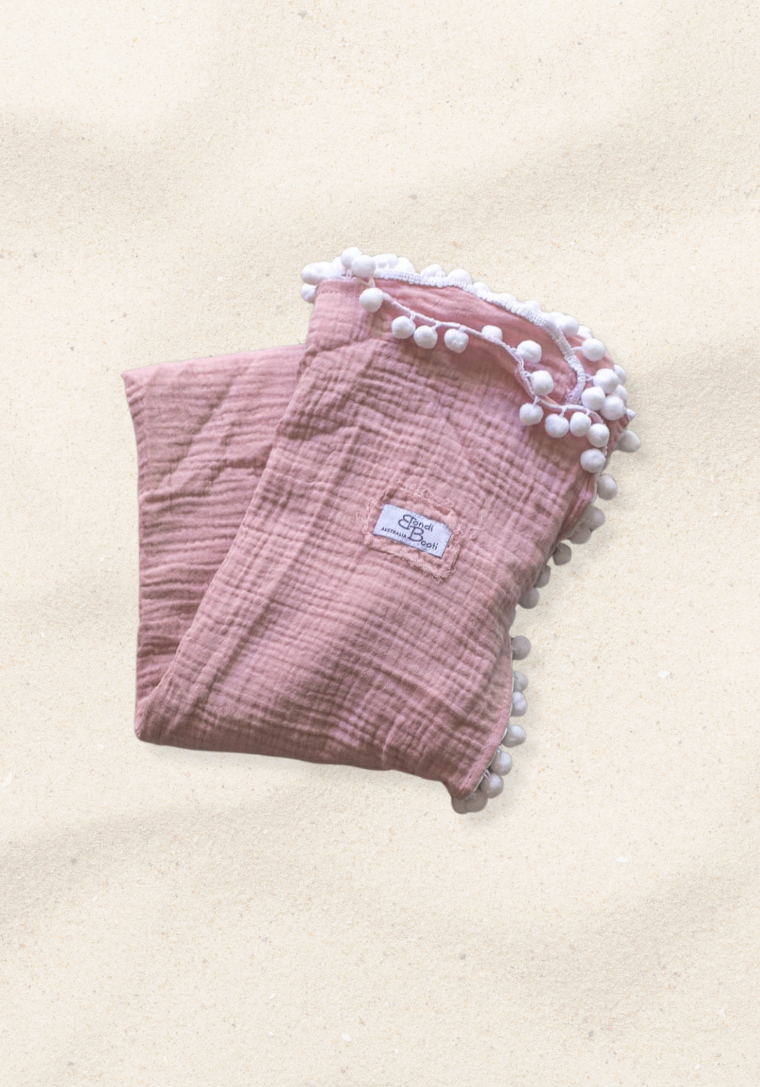 Dusty pink organic cotton baby blanket with soft white pom-pom trim, neatly folded with a visible Bondi Booti label, placed on a neutral background.