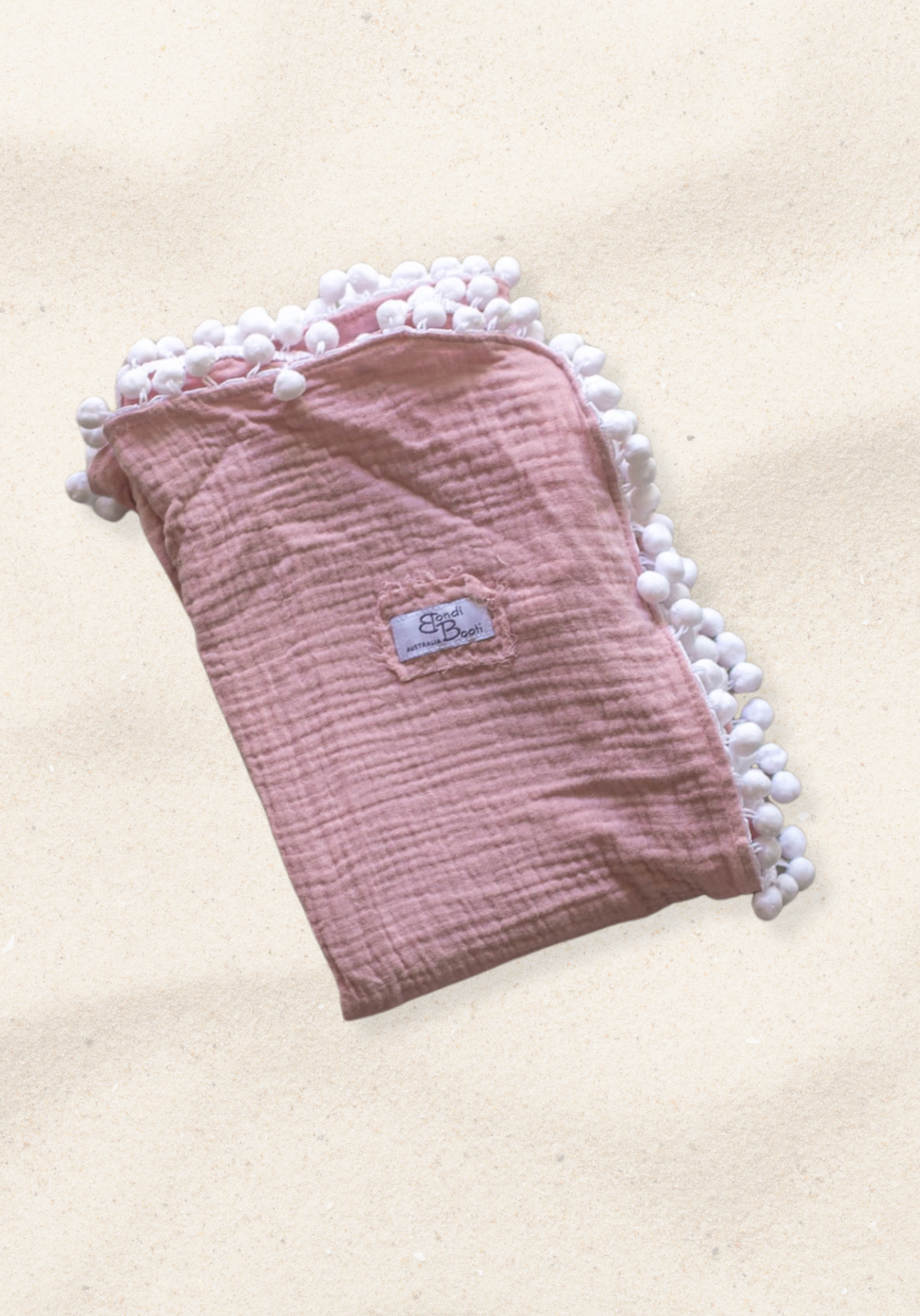 Dusty pink organic cotton baby blanket with soft white pom-pom trim, neatly folded with a visible Bondi Booti label, placed on a neutral background.
