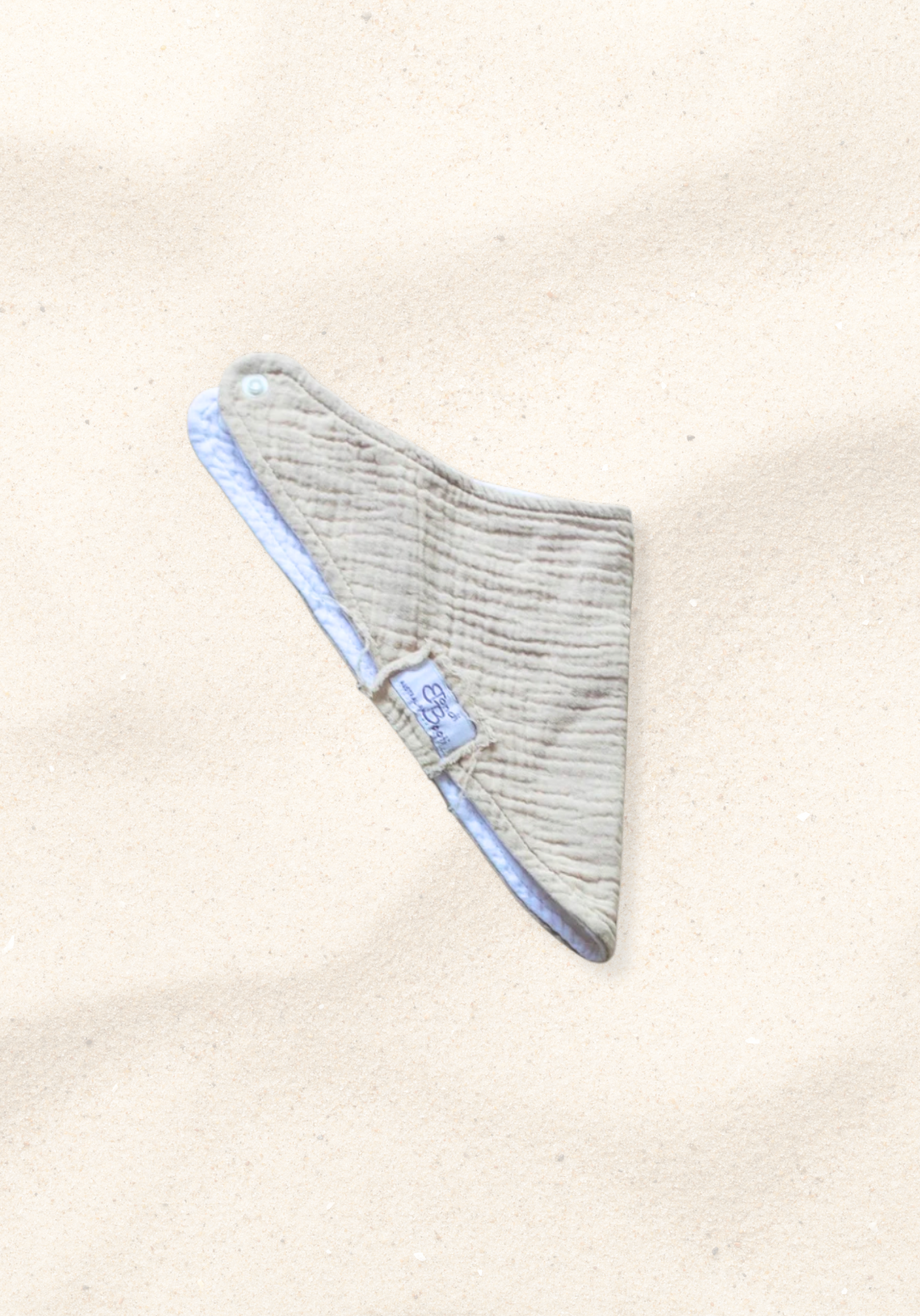 Soft beige organic cotton bandana dribble bib for babies with rounded shape and snap closure, displayed on a sand background
