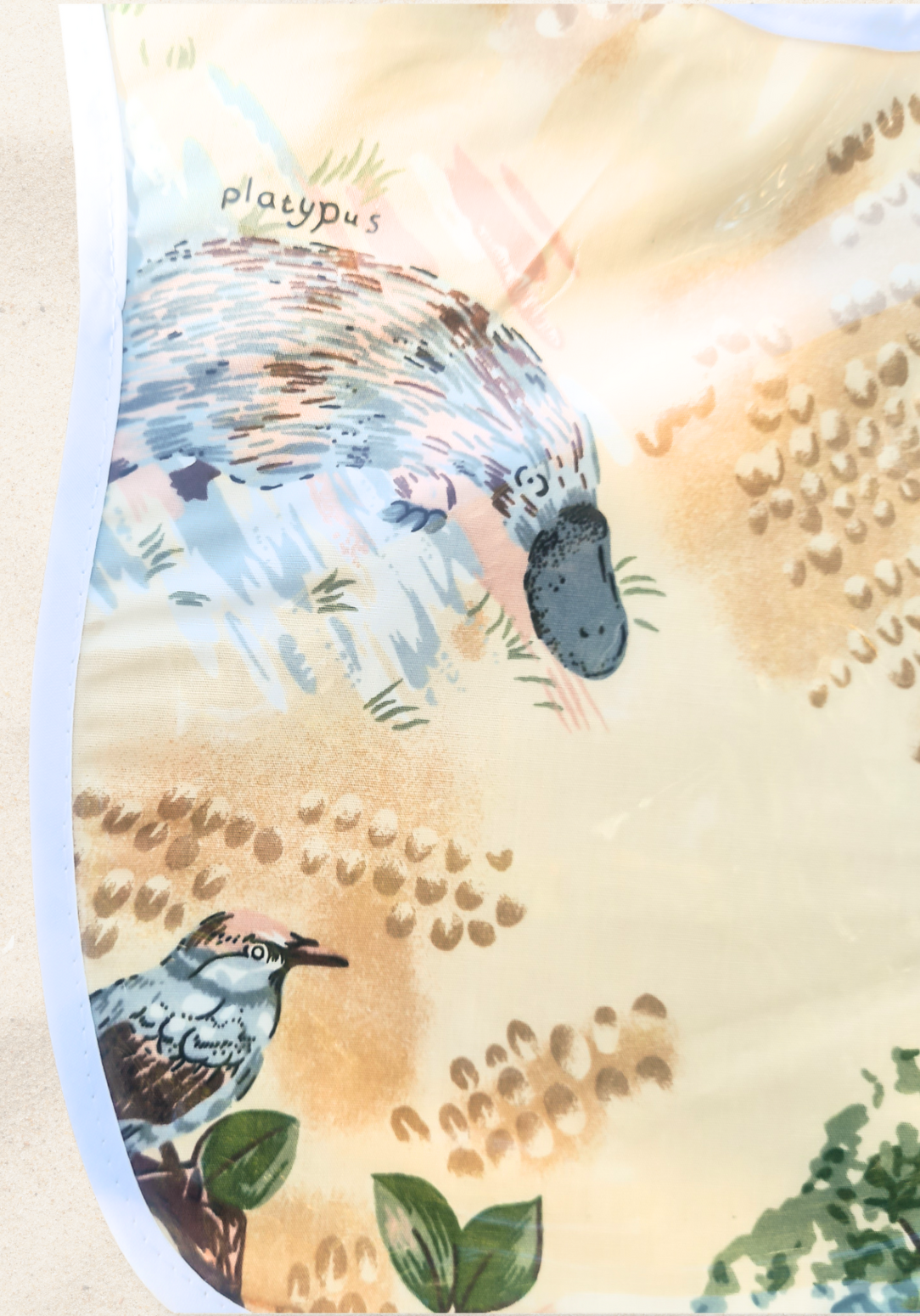 Laminated baby feeding bib featuring illustrated native Australian animals, including a platypus and birds, with a soft white trim and snap closure, laid flat on a sand background.