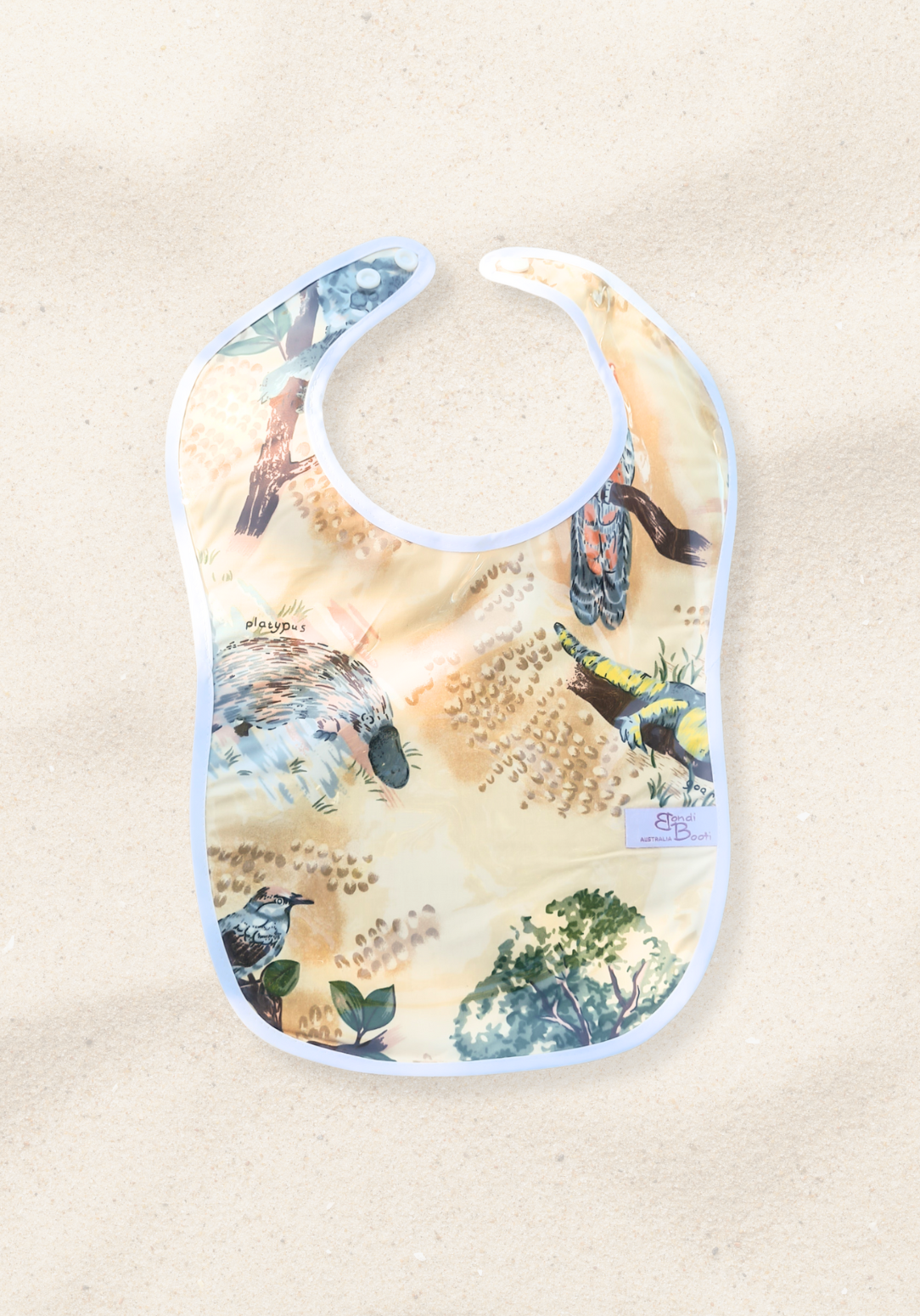 Laminated baby feeding bib featuring illustrated native Australian animals, including a platypus and birds, with a soft white trim and snap closure, laid flat on a sand background.
