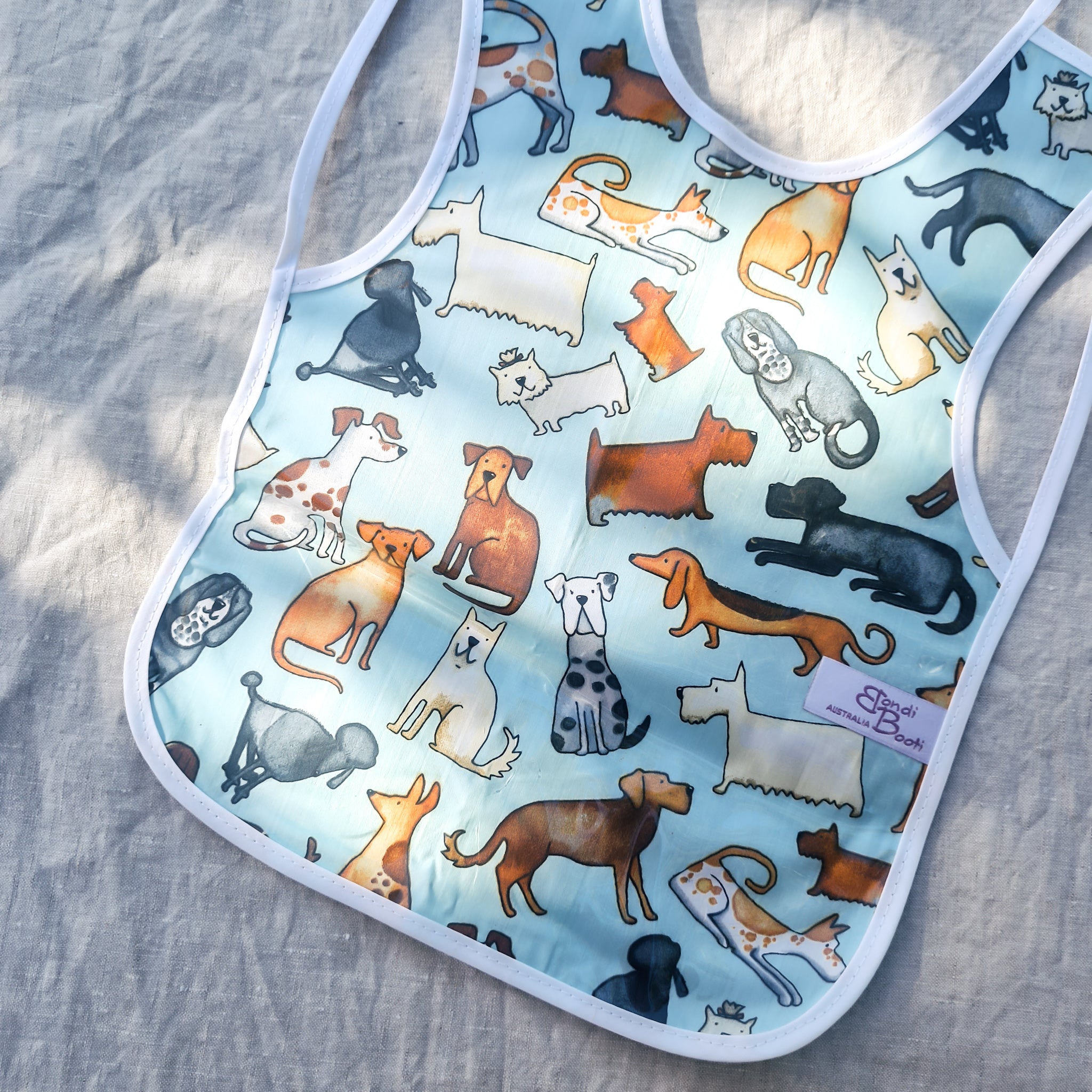 Laminated baby feeding bib featuring a playful pattern of illustrated dogs in various breeds and colors on a light blue background, trimmed in white with a snap closure, laid flat on a neutral fabric backdrop in soft natural light.