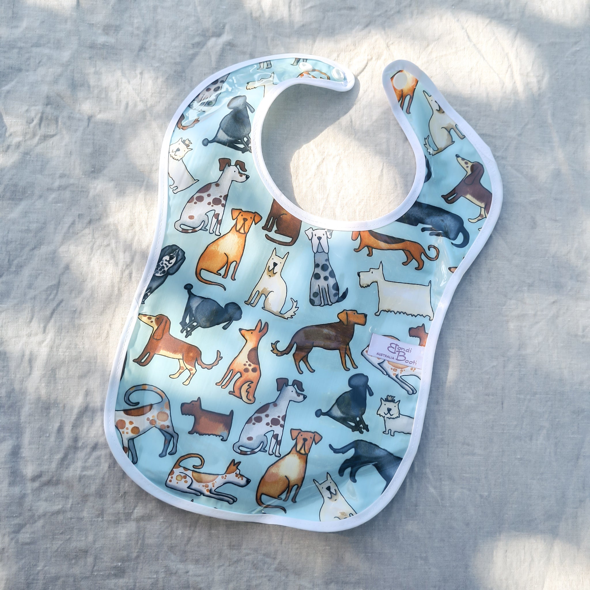 Laminated baby feeding bib featuring a playful pattern of illustrated dogs in various breeds and colors on a light blue background, trimmed in white with a snap closure, laid flat on a neutral fabric backdrop in soft natural light.