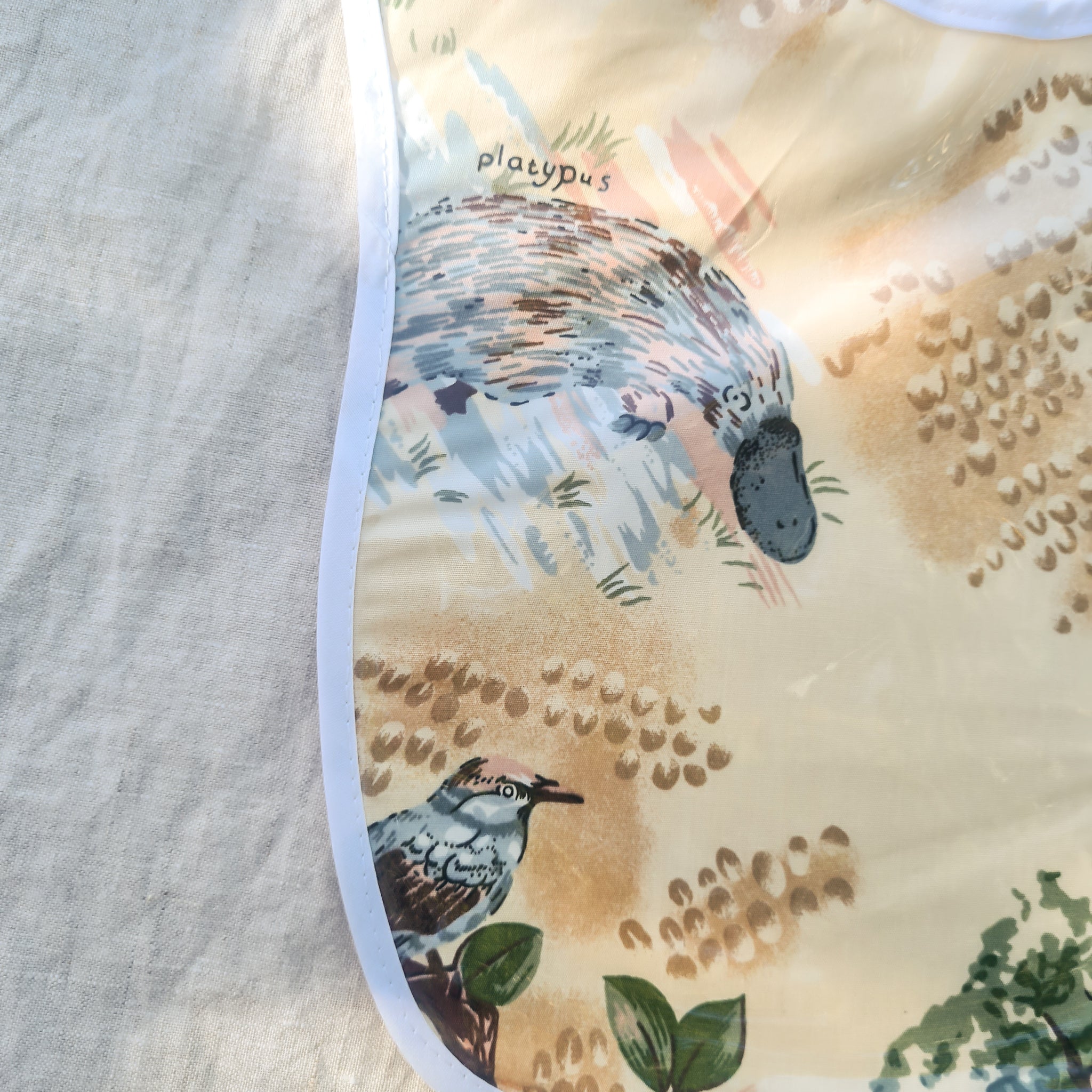 Laminated baby feeding bib featuring illustrated native Australian animals, including a platypus and birds, with a soft white trim and snap closure, laid flat on a neutral fabric background under dappled sunlight.