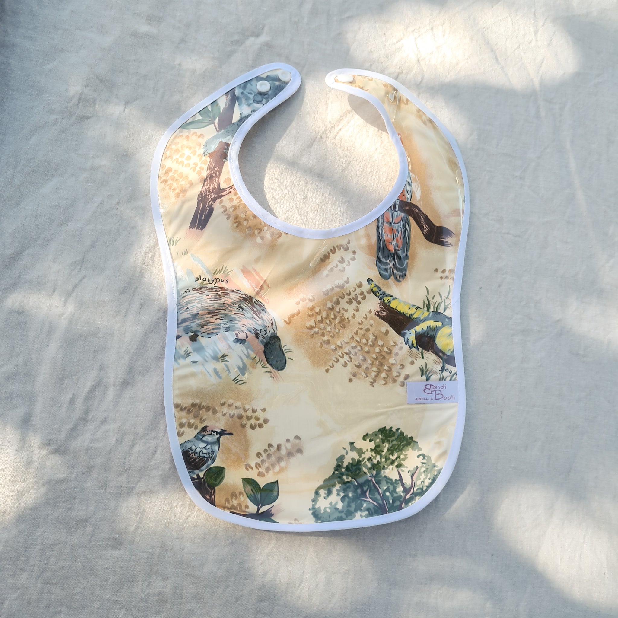 Laminated baby feeding bib featuring illustrated native Australian animals, including a platypus and birds, with a soft white trim and snap closure, laid flat on a neutral fabric background under dappled sunlight.